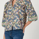 A woman wearing a short-sleeve button-front blouse with a dense paisley print in blue, green, brown, and beige tones, paired with blue jeans against a light gray studio background.