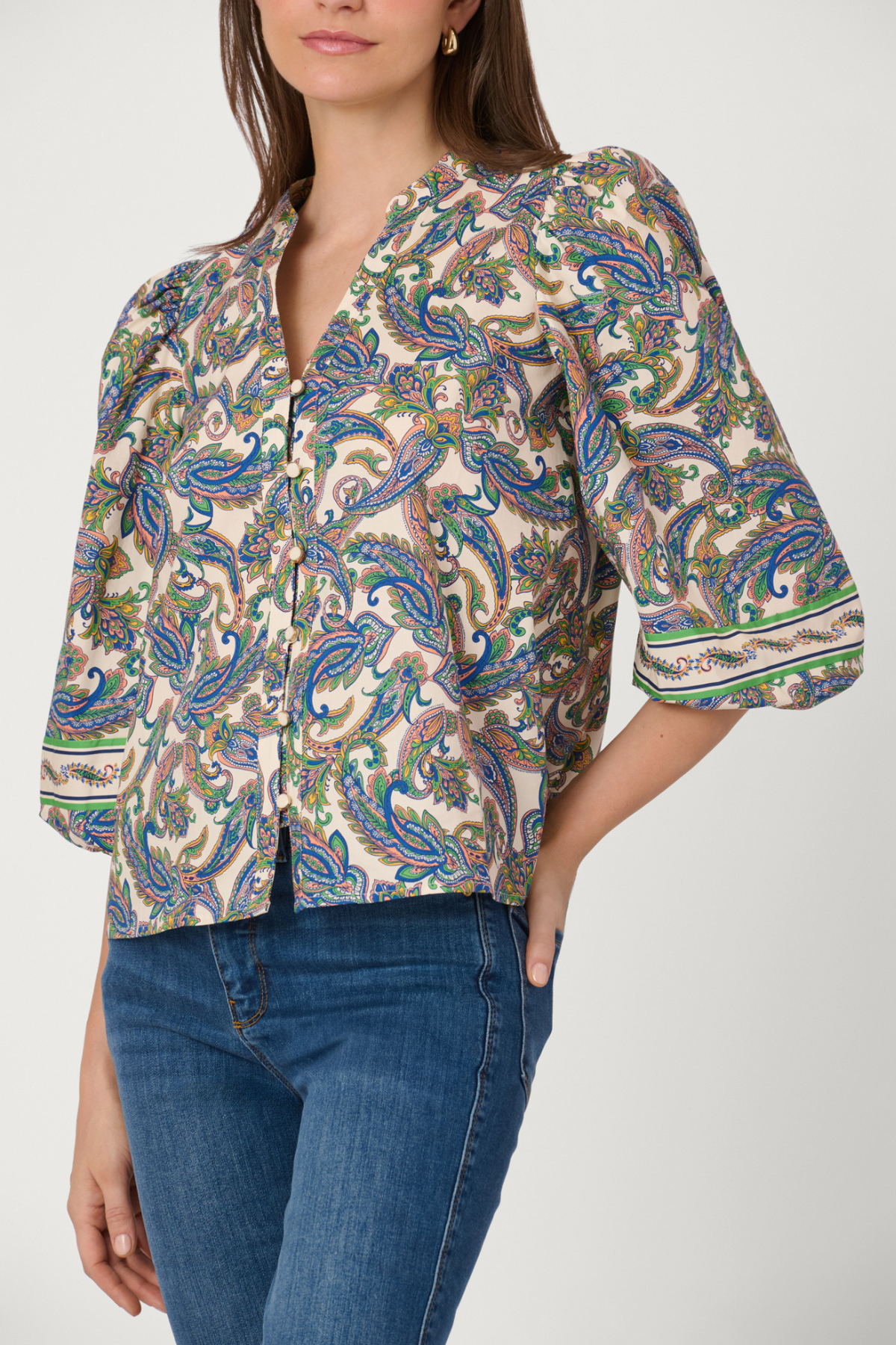A woman wearing a short-sleeve button-front blouse with a dense paisley print in blue, green, brown, and beige tones, paired with blue jeans against a light gray studio background.