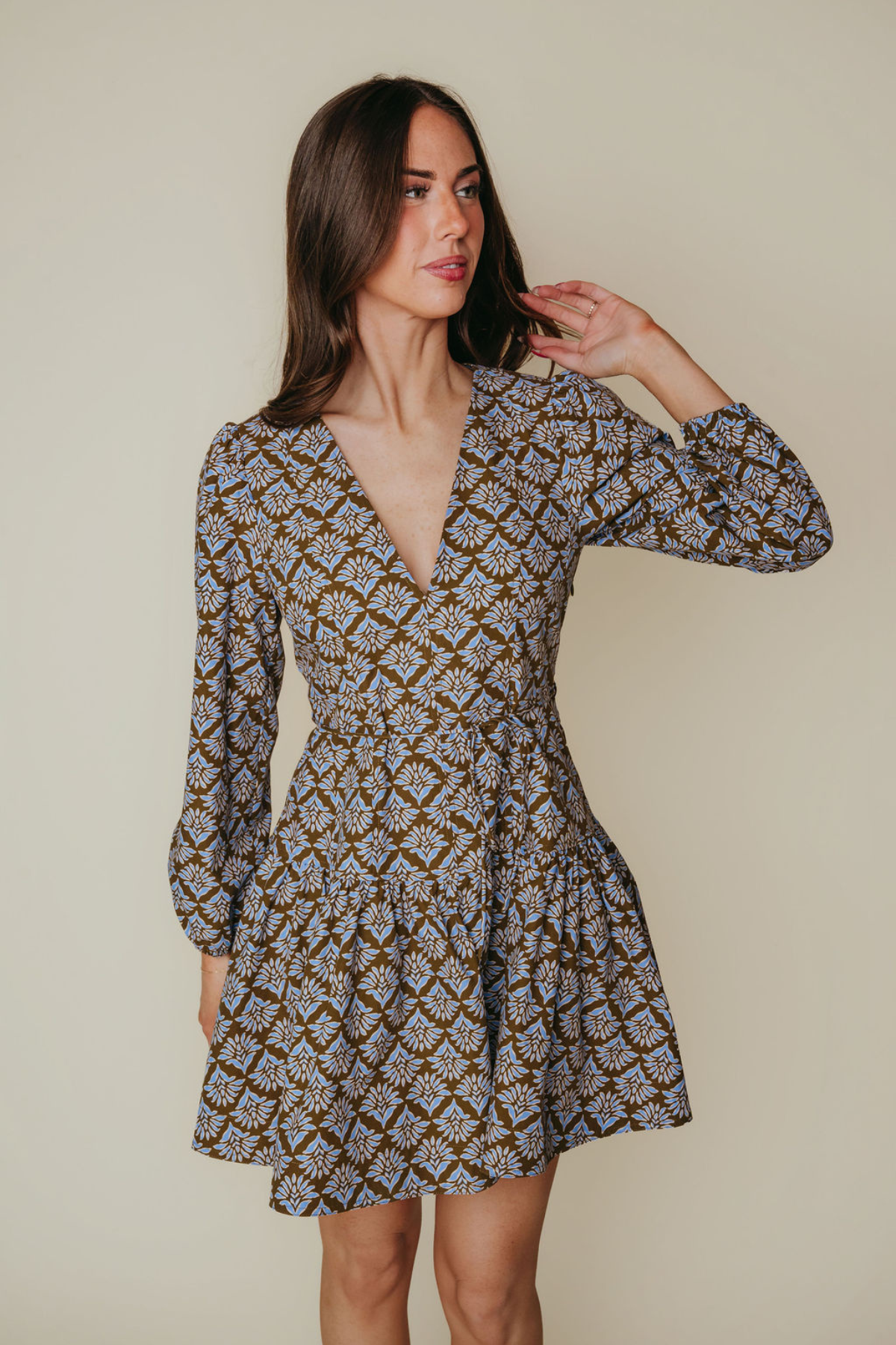 A woman modeling a long-sleeve mini dress with a brown, beige, and white geometric pattern against a solid beige background.