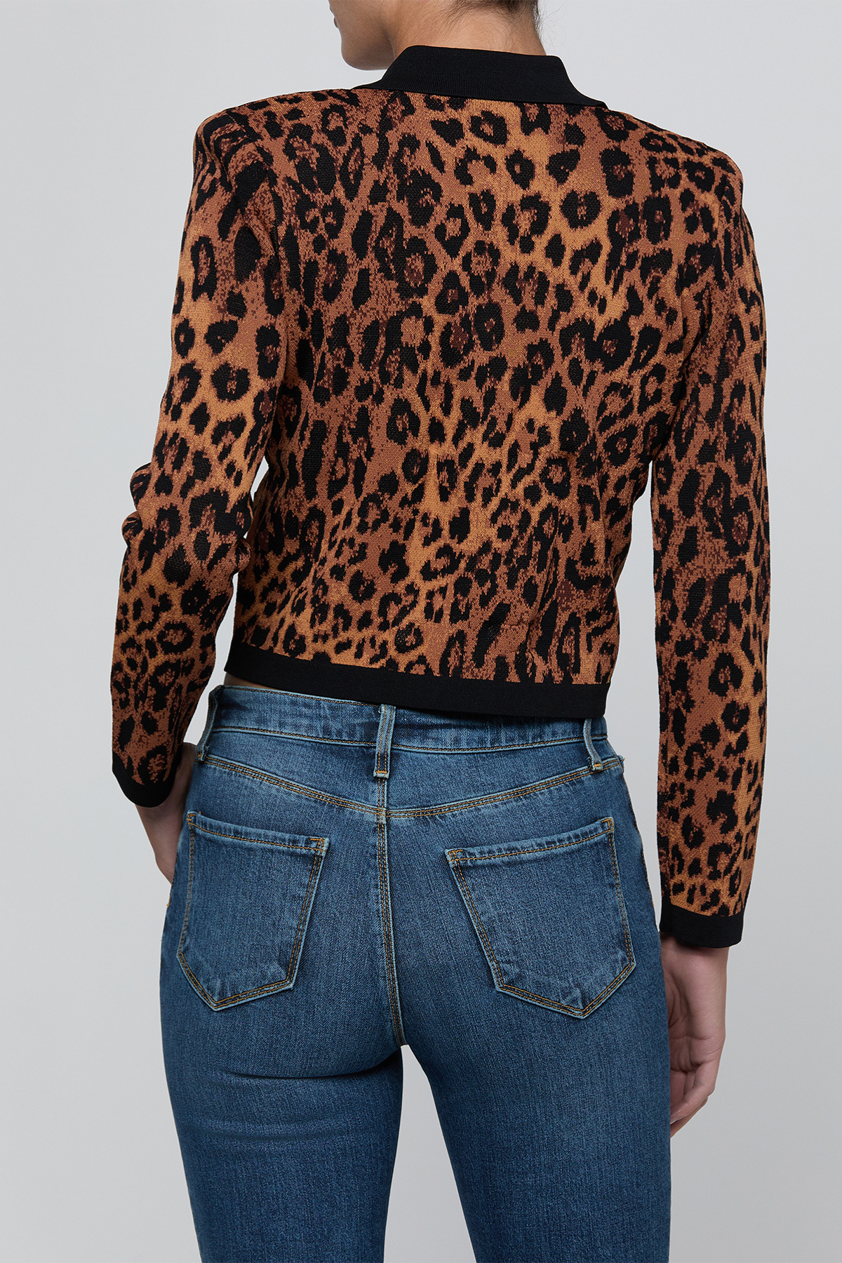 A person shown from the back wearing a cropped long-sleeve knit top with a brown and black leopard print, paired with blue jeans, against a light gray studio background.