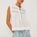 Woman wearing a white sleeveless top with blue embroidery and denim shorts on a white background