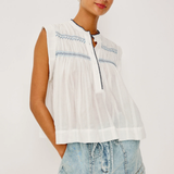 Woman wearing a white sleeveless top with blue embroidery and denim shorts on a white background