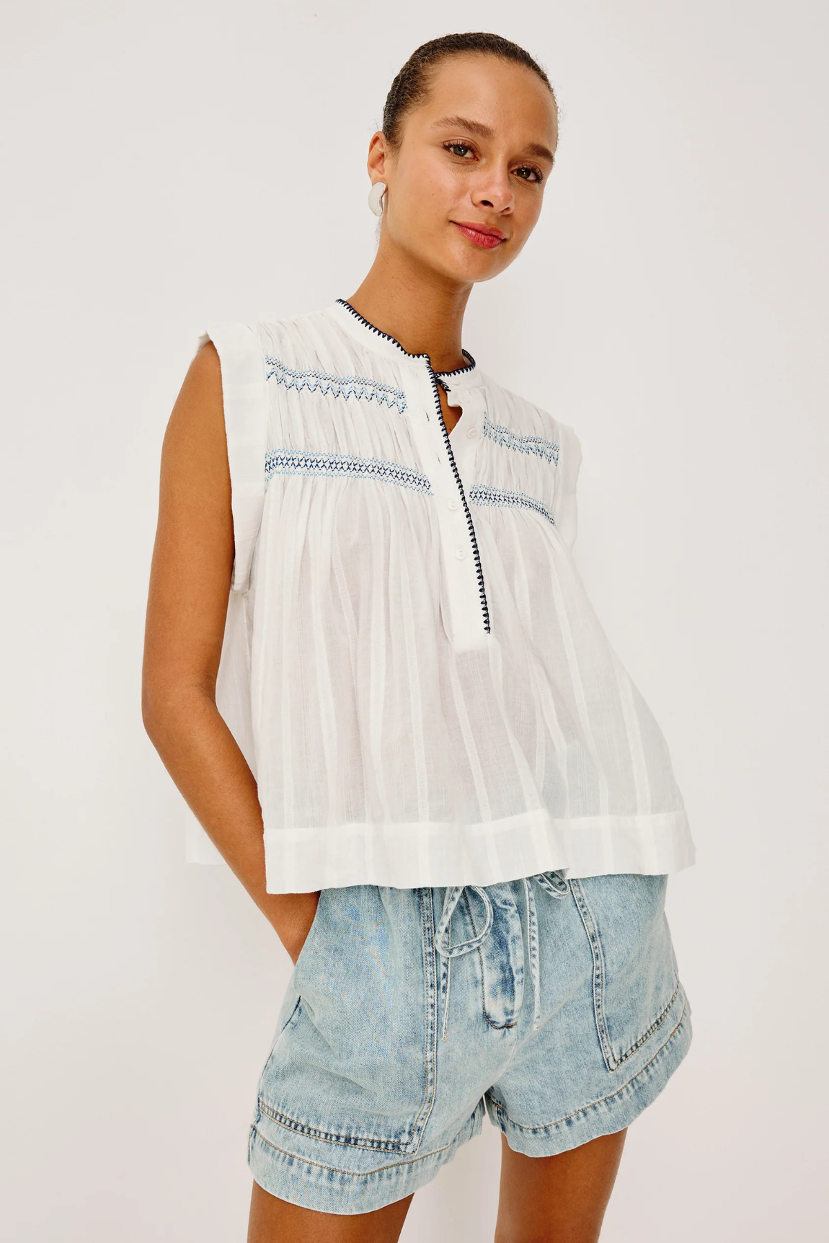 Woman wearing a white sleeveless top with blue embroidery and denim shorts on a white background