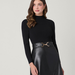 A woman modeling a black ribbed long-sleeve mock neck top paired with a black faux-leather mini skirt and a matching black belt with a gold buckle, set against a plain light gray studio background.