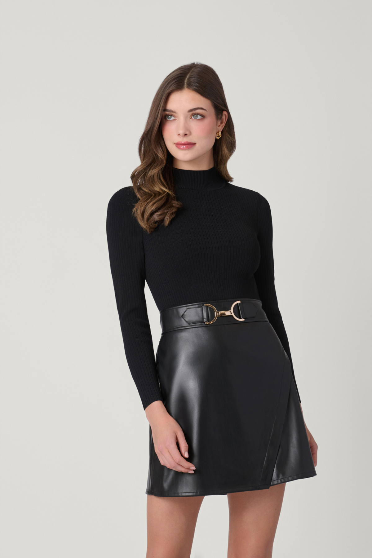 A woman modeling a black ribbed long-sleeve mock neck top paired with a black faux-leather mini skirt and a matching black belt with a gold buckle, set against a plain light gray studio background.