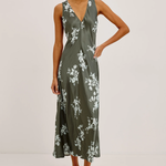 A woman modeling a sleeveless midi dress with a deep V-neckline and a white floral print on an olive green satin-like fabric, paired with white strappy high-heeled sandals against a plain light gray studio background.