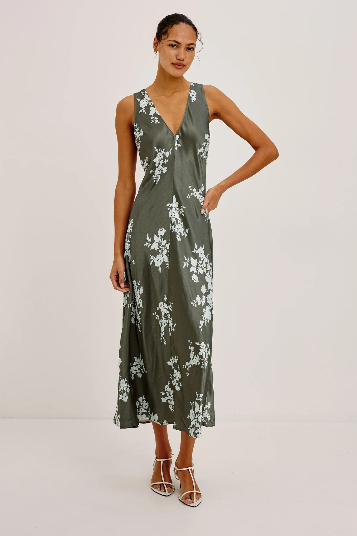 A woman modeling a sleeveless midi dress with a deep V-neckline and a white floral print on an olive green satin-like fabric, paired with white strappy high-heeled sandals against a plain light gray studio background.
