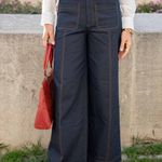 A person wearing dark navy wide-leg denim pants with contrast stitching, a white long-sleeve blouse, a tan leather belt, and tan woven flat shoes, holding a red handbag in an outdoor setting.