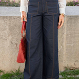 A person wearing dark navy wide-leg denim pants with contrast stitching, a white long-sleeve blouse, a tan leather belt, and tan woven flat shoes, holding a red handbag in an outdoor setting.