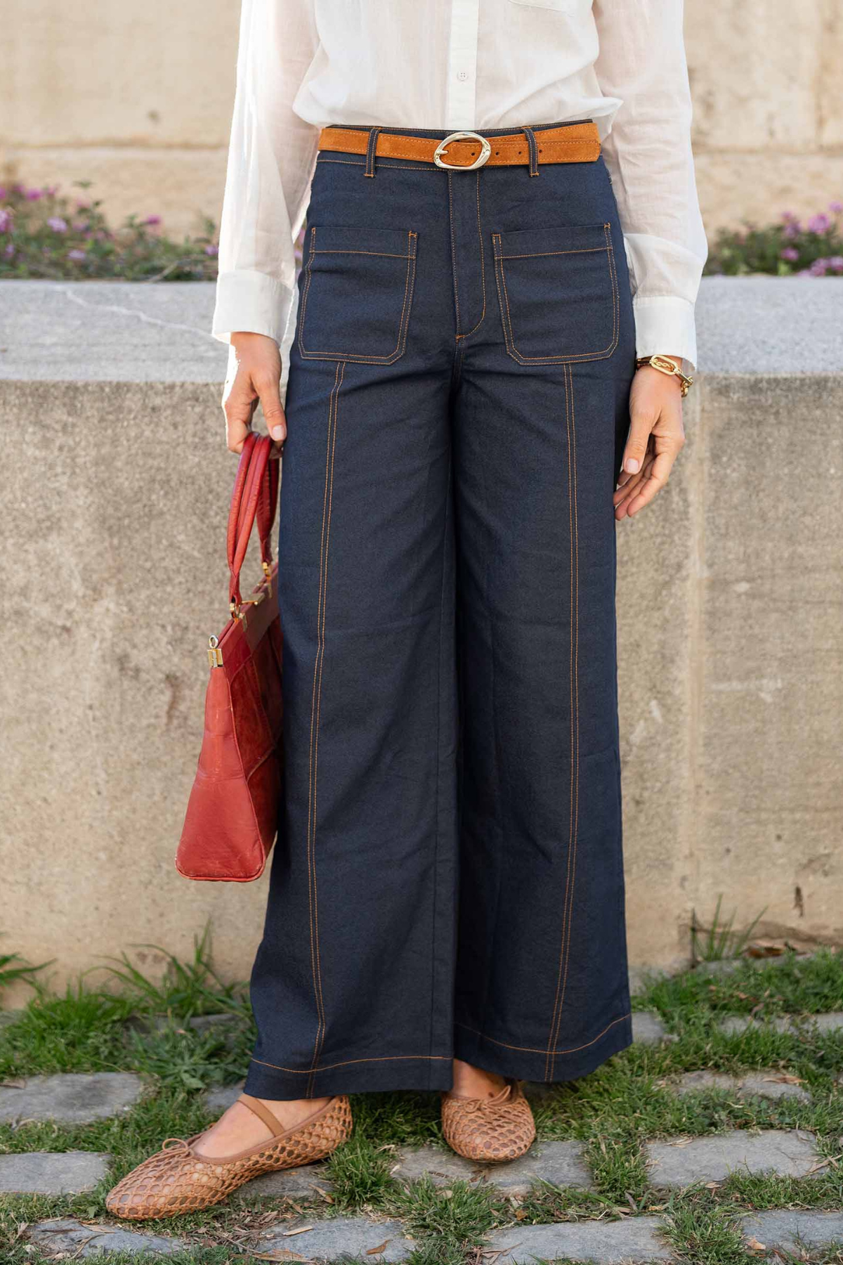 A person wearing dark navy wide-leg denim pants with contrast stitching, a white long-sleeve blouse, a tan leather belt, and tan woven flat shoes, holding a red handbag in an outdoor setting.