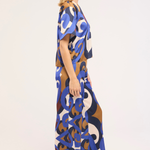 A woman shown in side profile wearing a loose, floor-length kaftan dress with an abstract, interlocking loop pattern in blue, brown, cream, and navy on a white background.
