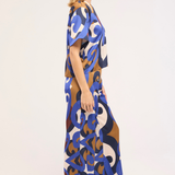 A woman shown in side profile wearing a loose, floor-length kaftan dress with an abstract, interlocking loop pattern in blue, brown, cream, and navy on a white background.