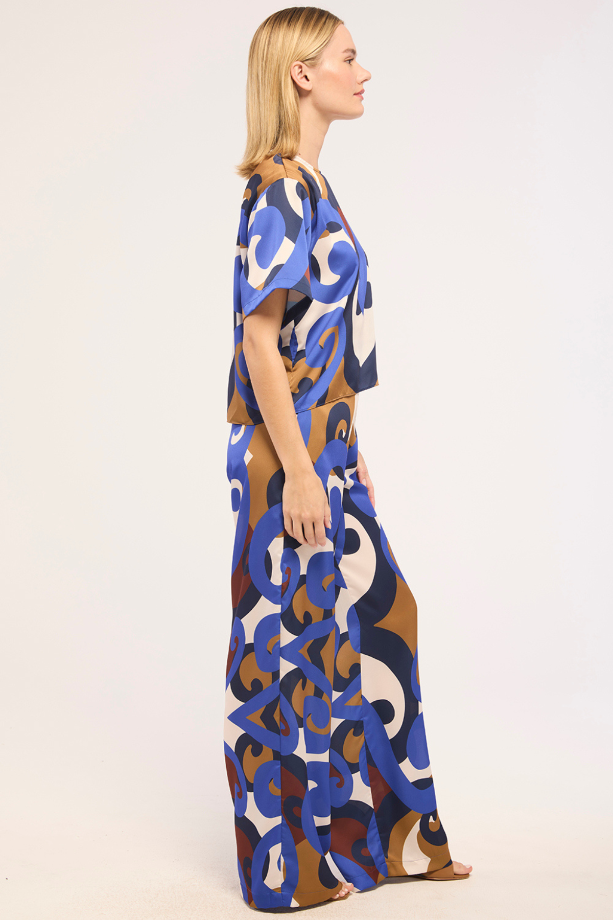 A woman shown in side profile wearing a loose, floor-length kaftan dress with an abstract, interlocking loop pattern in blue, brown, cream, and navy on a white background.