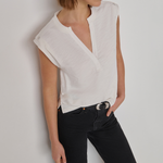 A woman wearing a white sleeveless wrap-style blouse with a split neckline and black jeans, standing against a plain light gray wall.