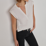 A woman wearing a white sleeveless wrap-style blouse with a split neckline and black jeans, standing against a plain light gray wall.