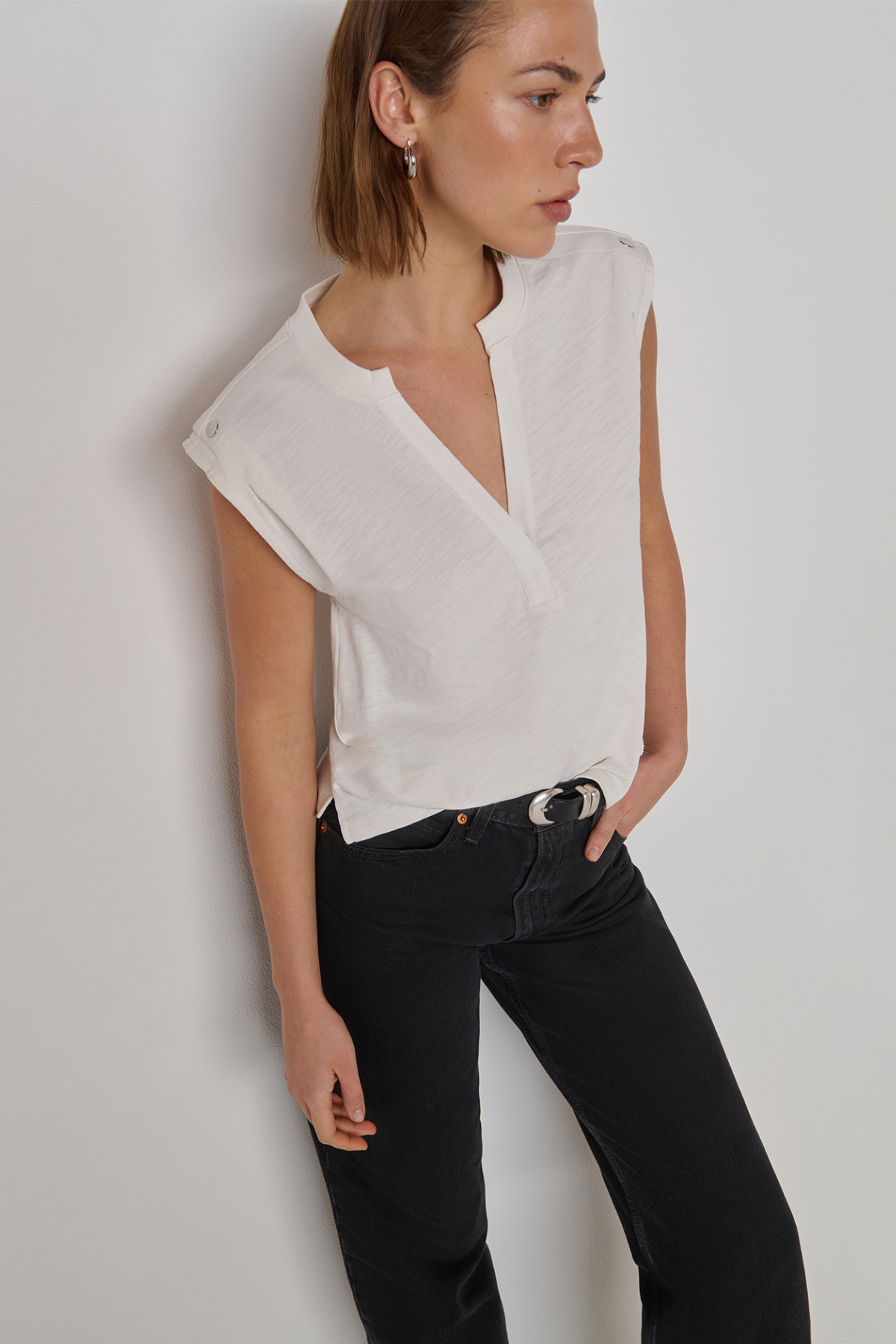 A woman wearing a white sleeveless wrap-style blouse with a split neckline and black jeans, standing against a plain light gray wall.