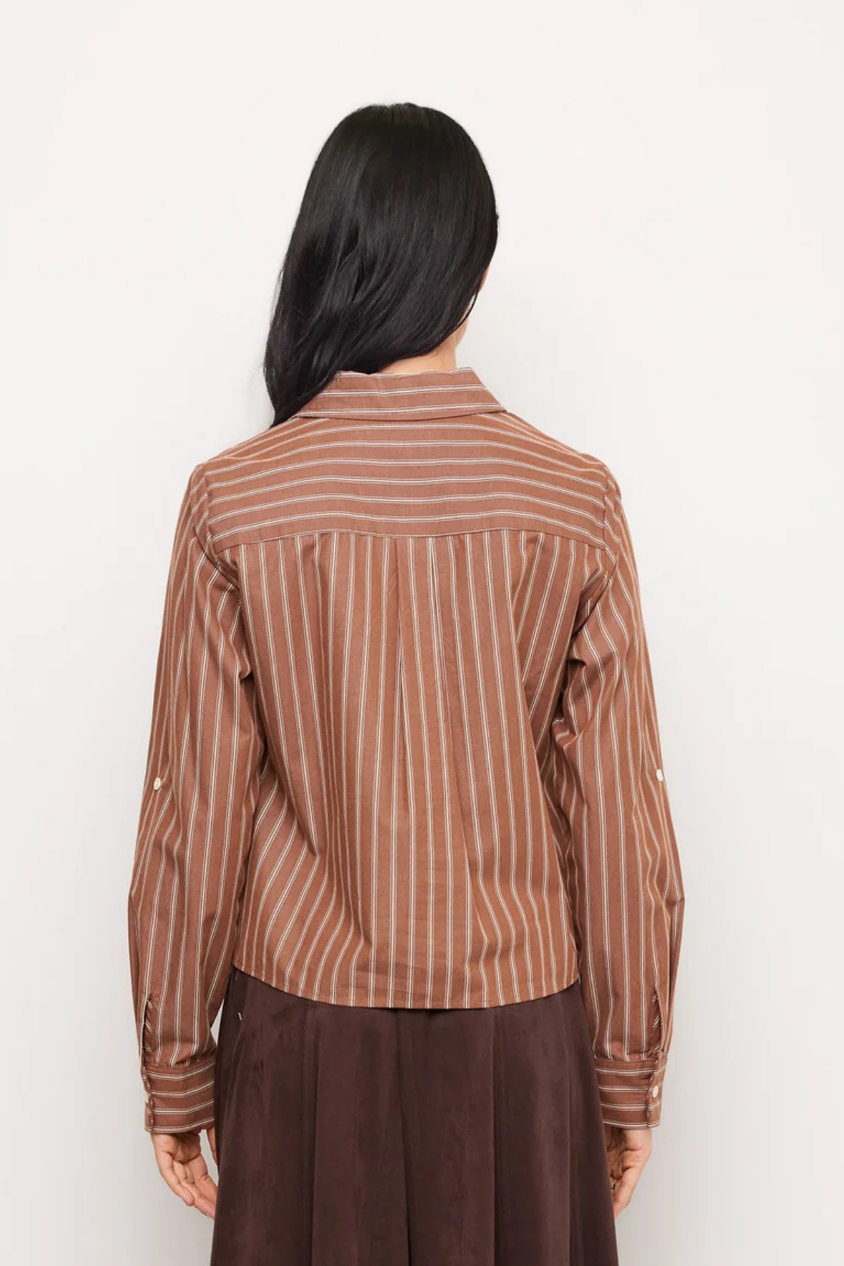 A woman viewed from behind wearing a brown and beige vertically striped long-sleeve shirt and a dark brown skirt, set against a plain white background.
