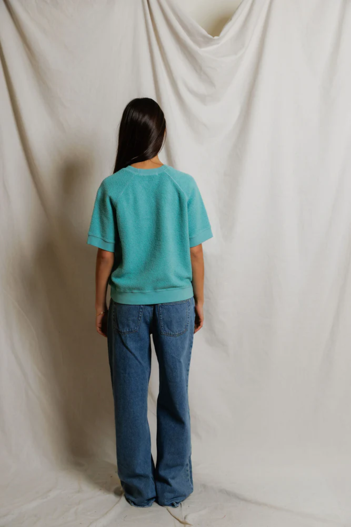 Person wearing a teal shirt and blue jeans standing against a plain white background