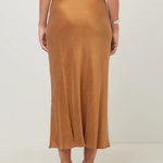 A woman is shown from the back wearing a flowing midi skirt in camel brown, paired with a navy top and black heels, against a plain light gray studio background.