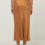 A woman is shown from the back wearing a flowing midi skirt in camel brown, paired with a navy top and black heels, against a plain light gray studio background.