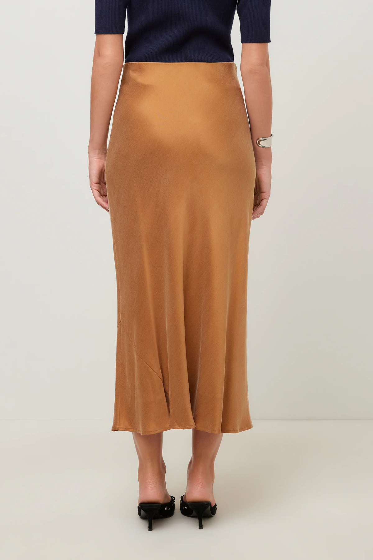 A woman is shown from the back wearing a flowing midi skirt in camel brown, paired with a navy top and black heels, against a plain light gray studio background.