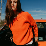 Person wearing an orange sweater standing next to a red car with a clear blue sky.