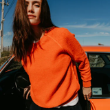 Person wearing an orange sweater standing next to a red car with a clear blue sky.