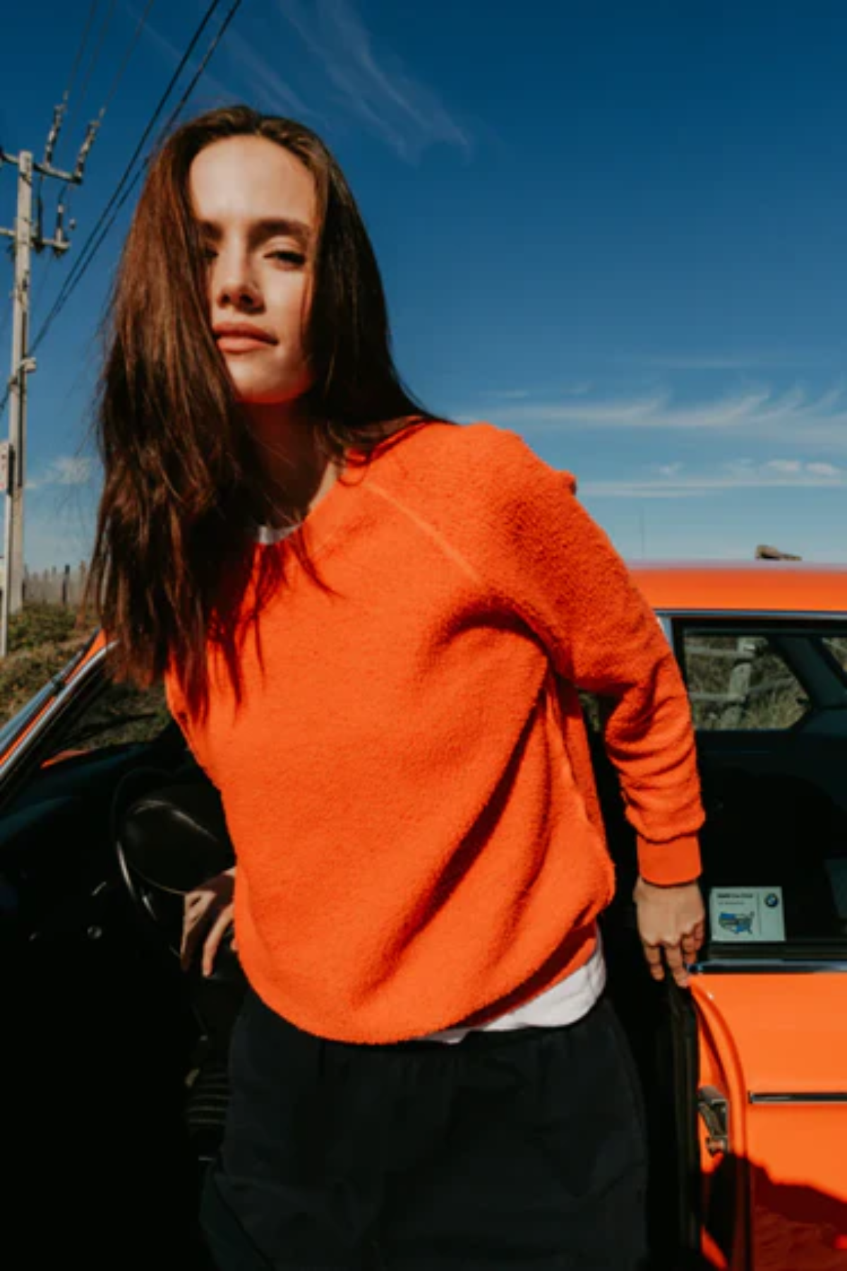 Person wearing an orange sweater standing next to a red car with a clear blue sky.