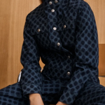 A model wearing a dark navy blue patterned jumpsuit with silver buttons, seated against a light wood-paneled background.