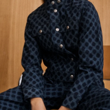 A model wearing a dark navy blue patterned jumpsuit with silver buttons, seated against a light wood-paneled background.