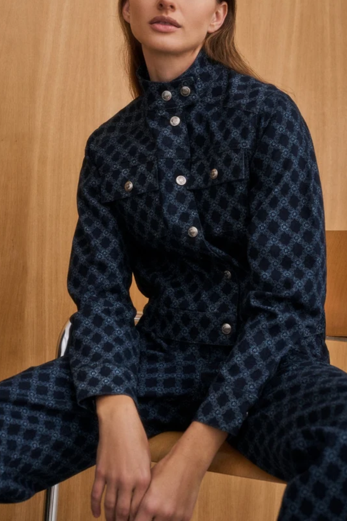 A model wearing a dark navy blue patterned jumpsuit with silver buttons, seated against a light wood-paneled background.
