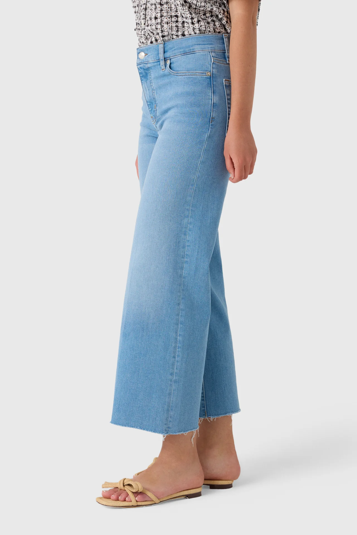 A model wearing light blue cropped flare jeans with a frayed hem, paired with flat slide sandals, shown from the waist down on a light gray studio background.