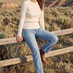 A woman stands outdoors in a desert canyon landscape, leaning against a wooden fence. She is wearing a cream ribbed knit sweater, light blue jeans, and brown ankle boots.