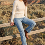 A woman stands outdoors in a desert canyon landscape, leaning against a wooden fence. She is wearing a cream ribbed knit sweater, light blue jeans, and brown ankle boots.