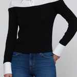 A woman wearing a black off-the-shoulder knit sweater with contrasting white collar and cuffs, paired with blue jeans, against a light gray studio background.