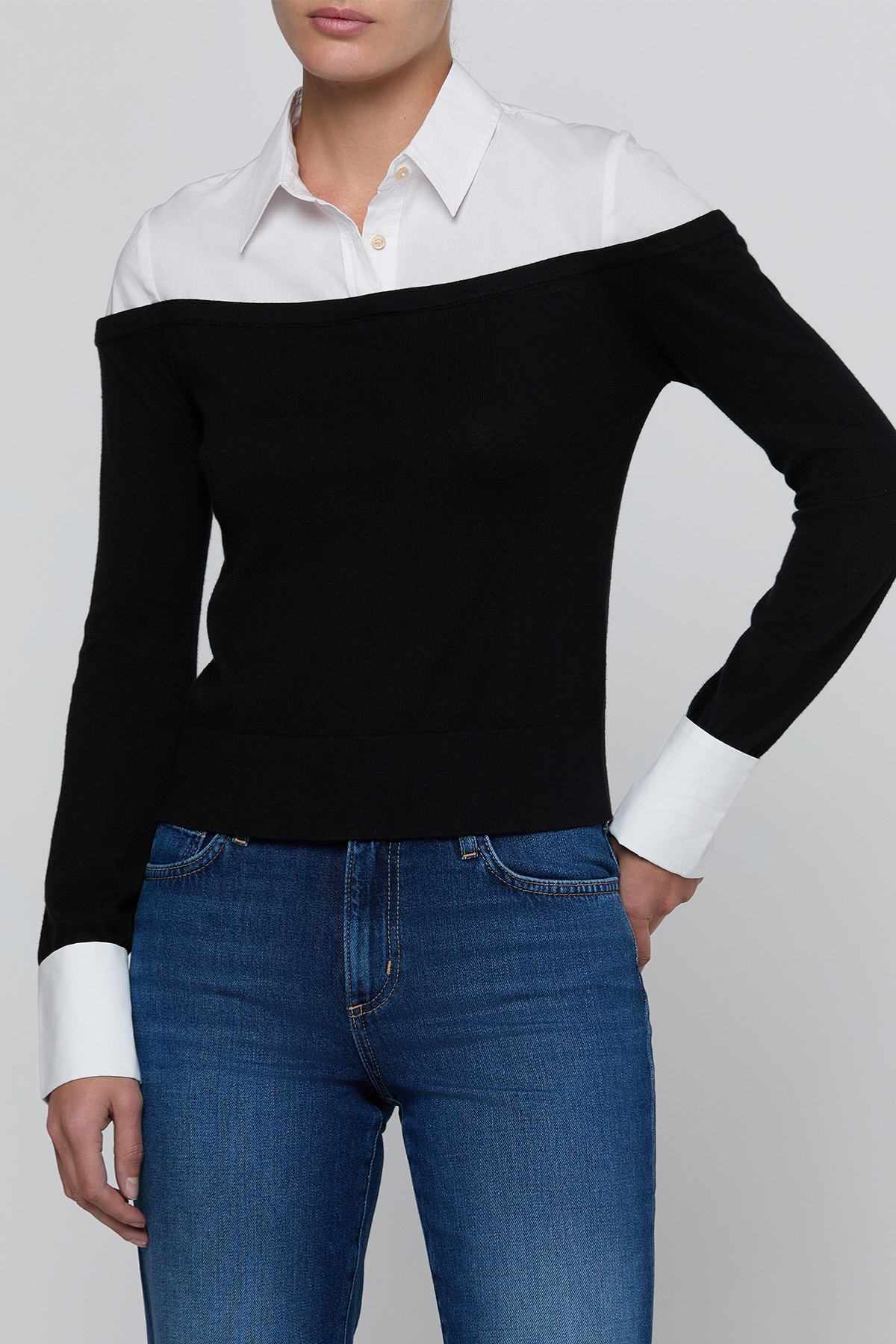 A woman wearing a black off-the-shoulder knit sweater with contrasting white collar and cuffs, paired with blue jeans, against a light gray studio background.