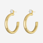 A pair of gold hoop earrings with a small clear round gemstone at the top of each earring, shown on a light gray background.