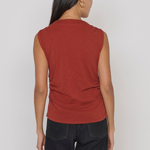 A woman shown from the back wearing a sleeveless rust-red top and dark jeans against a plain light gray studio background.
