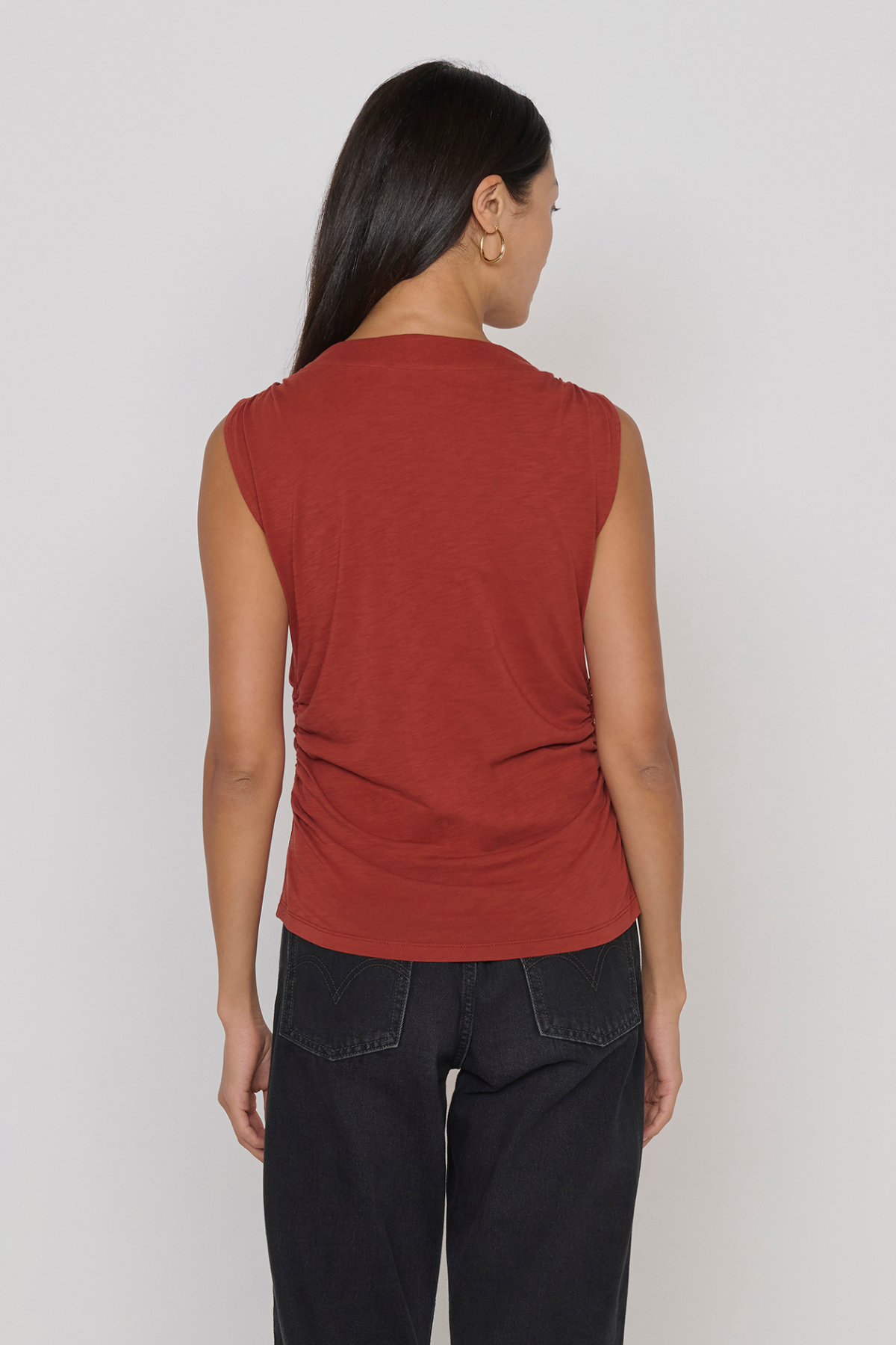 A woman shown from the back wearing a sleeveless rust-red top and dark jeans against a plain light gray studio background.