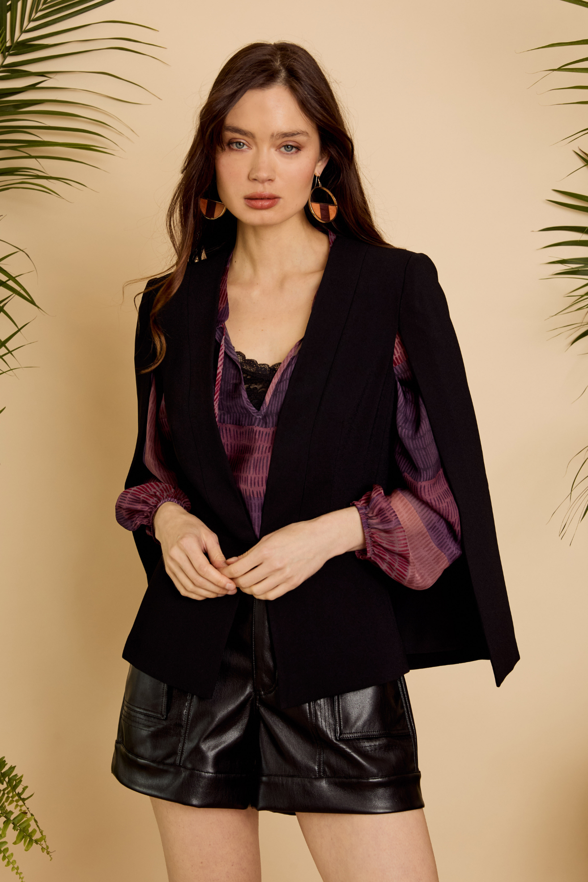 Woman wearing a black blazer with purple accents and leather shorts against a beige background