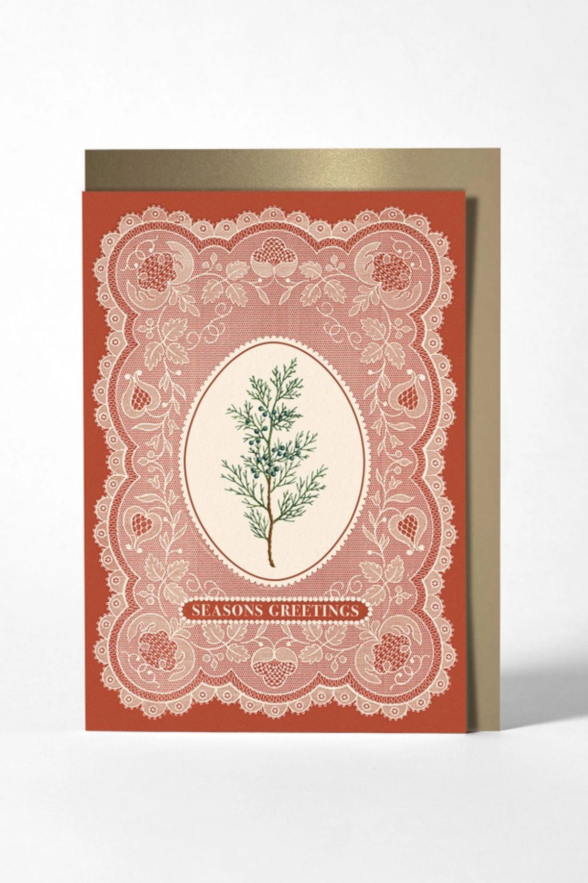 A vintage-style holiday greeting card with a terracotta red border, white lace-like ornamental pattern, and a central cream oval featuring a green pine branch. The card reads 'SEASONS GREETINGS' and is shown standing upright with a metallic gold envelope behind it on a light gray background.