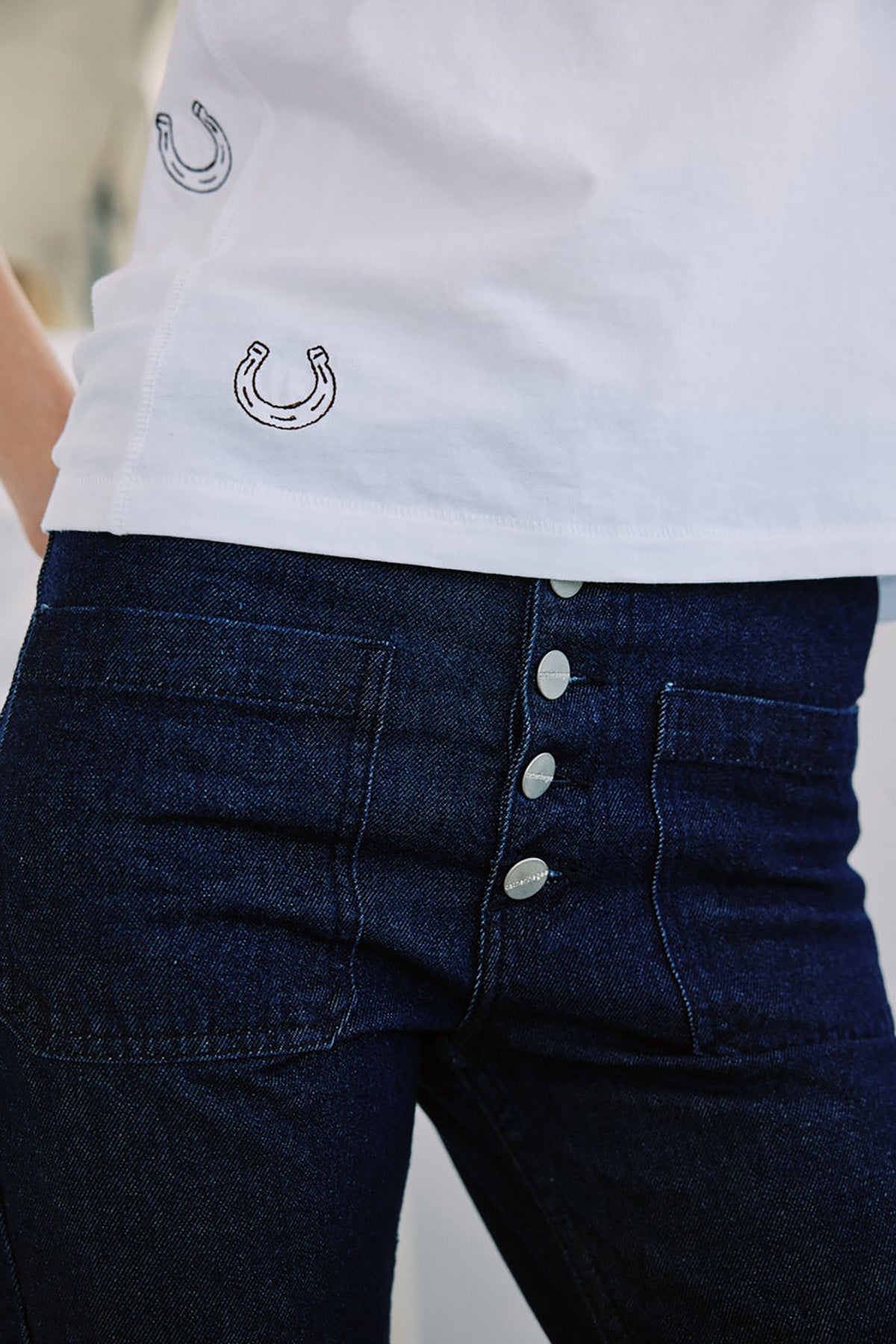 White shirt with embroidered horseshoe design and dark blue jeans with silver buttons.