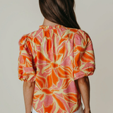Woman wearing a colorful floral blouse with white pants against a plain background