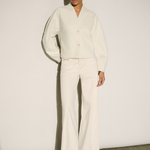 A model stands against a minimalist off-white wall wearing a matching cream-colored two-piece set consisting of a cropped long-sleeve button-up blouse with balloon sleeves and matching wide-leg trousers, paired with beige pointed-toe heels.