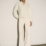 A model stands against a minimalist off-white wall wearing a matching cream-colored two-piece set consisting of a cropped long-sleeve button-up blouse with balloon sleeves and matching wide-leg trousers, paired with beige pointed-toe heels.