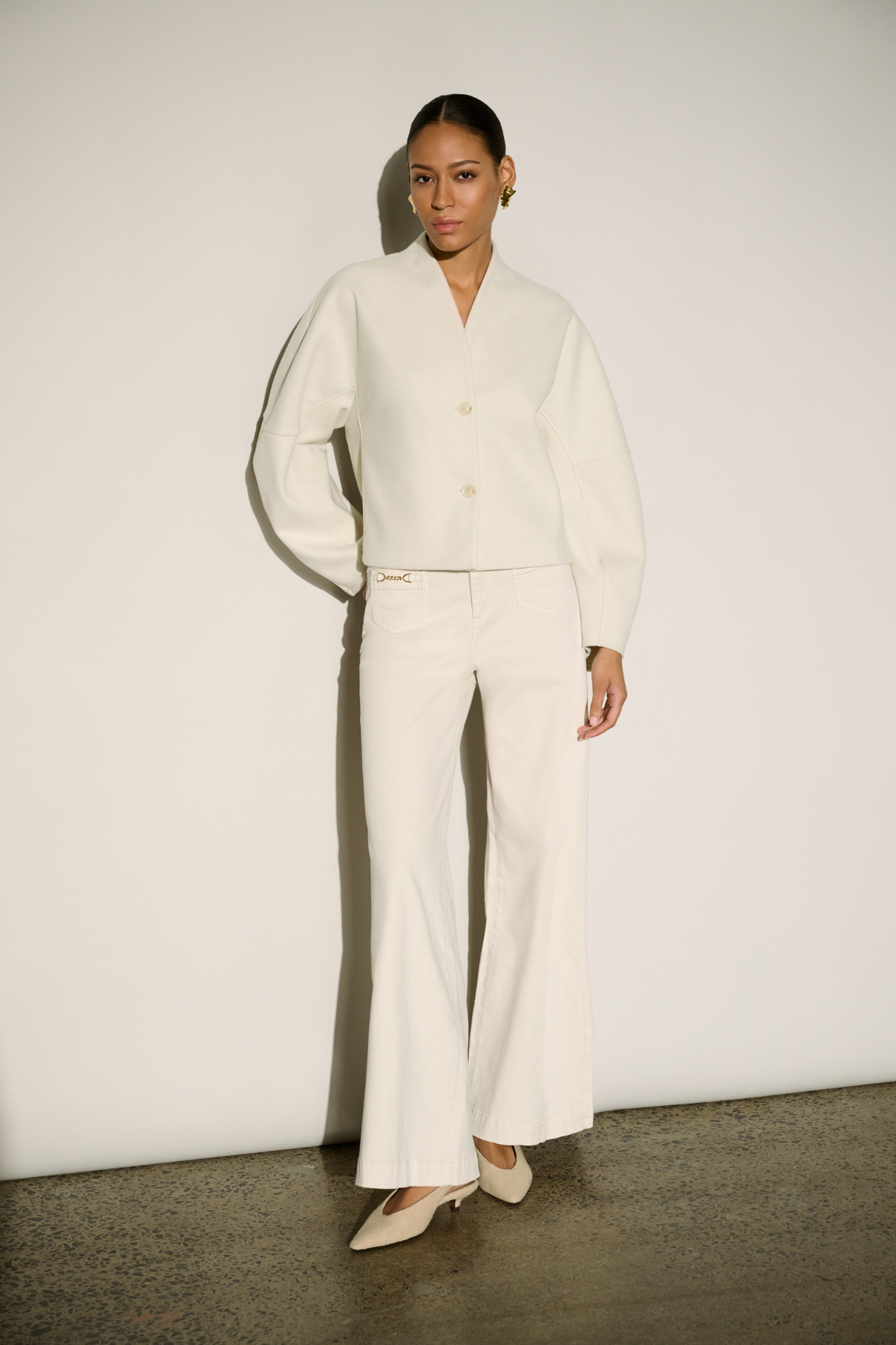 A model stands against a minimalist off-white wall wearing a matching cream-colored two-piece set consisting of a cropped long-sleeve button-up blouse with balloon sleeves and matching wide-leg trousers, paired with beige pointed-toe heels.