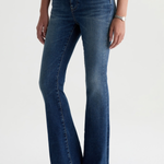 A model wearing a pair of dark blue flare jeans with a black top and black pointed-toe heels, shown from the waist down against a light gray studio background.