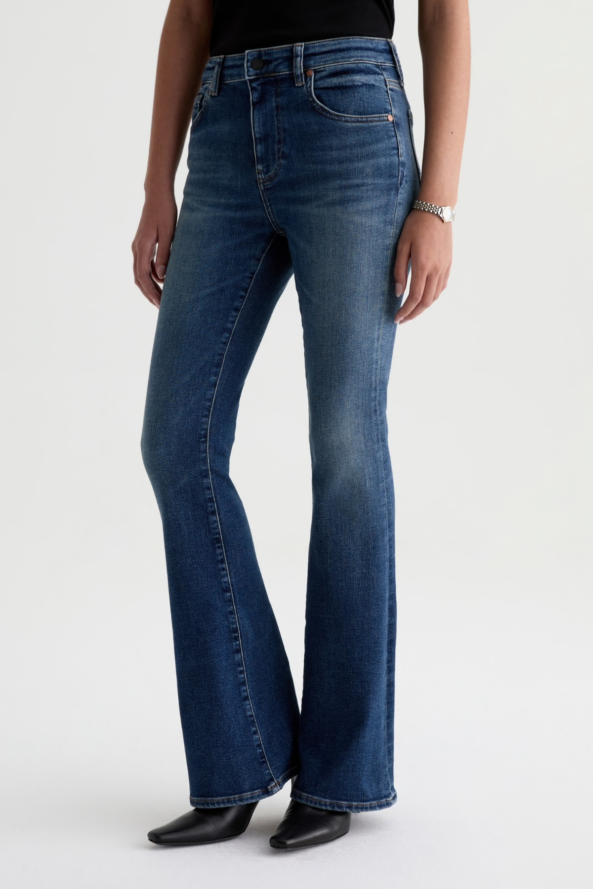 A model wearing a pair of dark blue flare jeans with a black top and black pointed-toe heels, shown from the waist down against a light gray studio background.