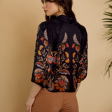 A woman shown from the back wearing a dark navy floral blouse with orange, red, and beige botanical embroidery, paired with brown pants, against a warm beige background with plant leaves at the edges.