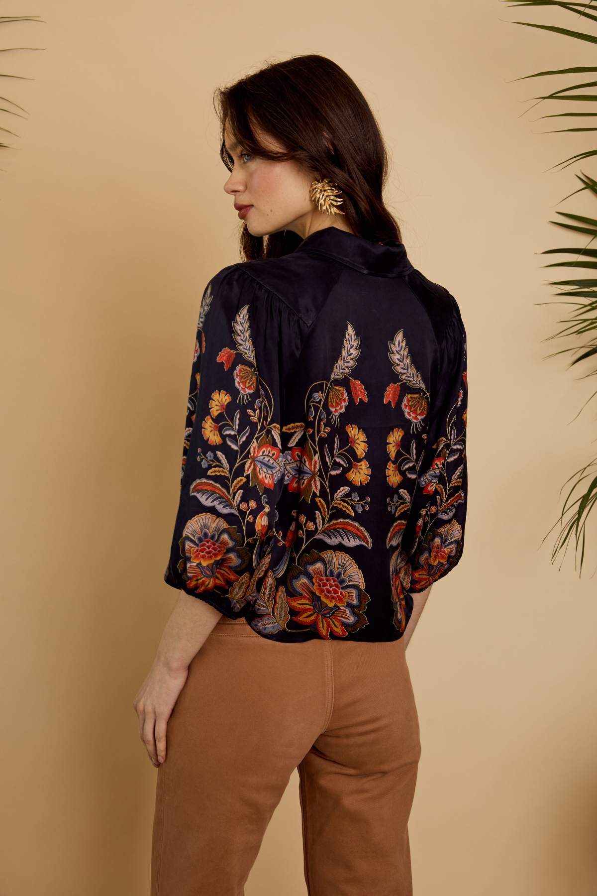 A woman shown from the back wearing a dark navy floral blouse with orange, red, and beige botanical embroidery, paired with brown pants, against a warm beige background with plant leaves at the edges.